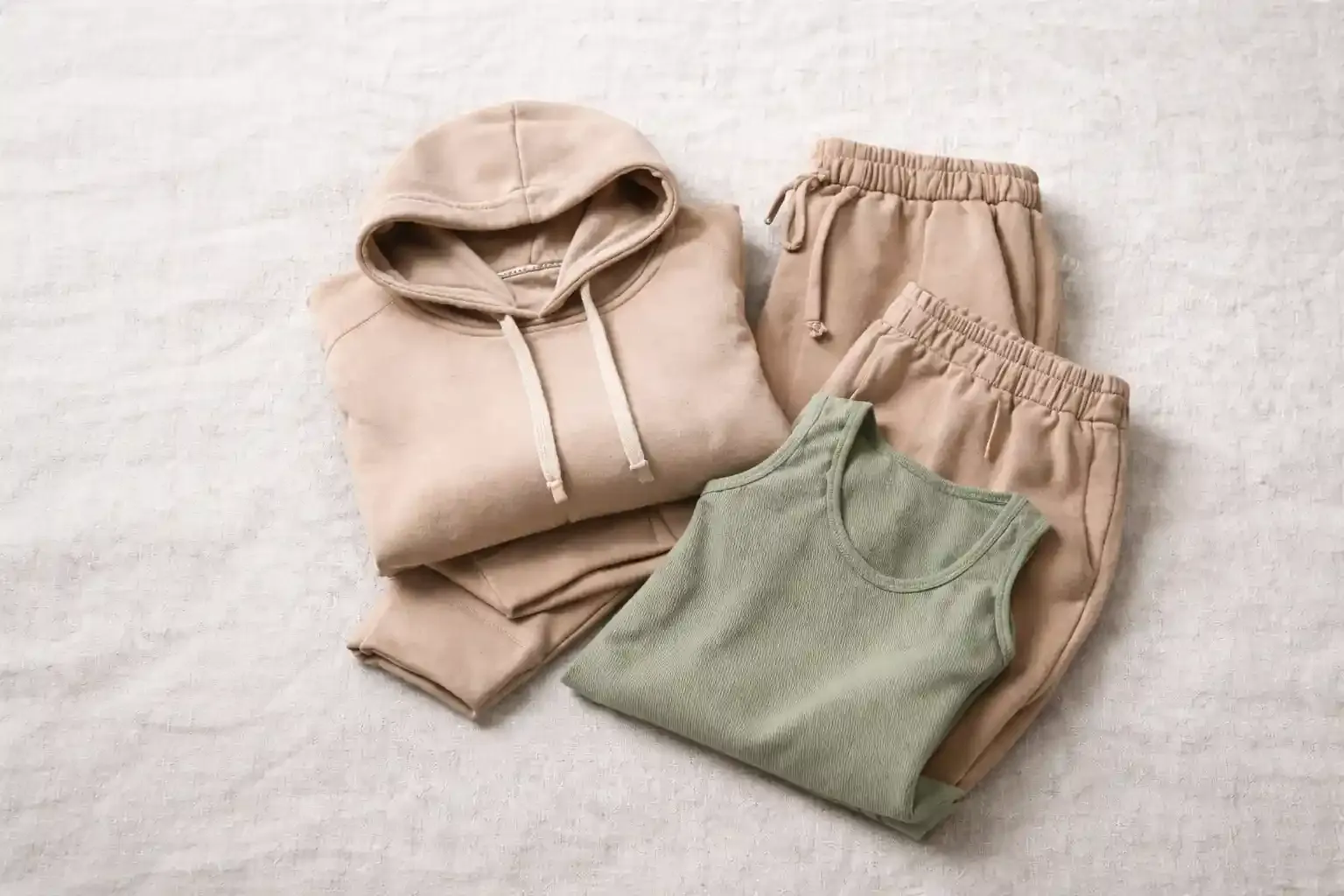 Private label loungewear set with beige hoodie, matching sweatpants, and sage green ribbed tank on light textured background, showing fabric quality and soft texture